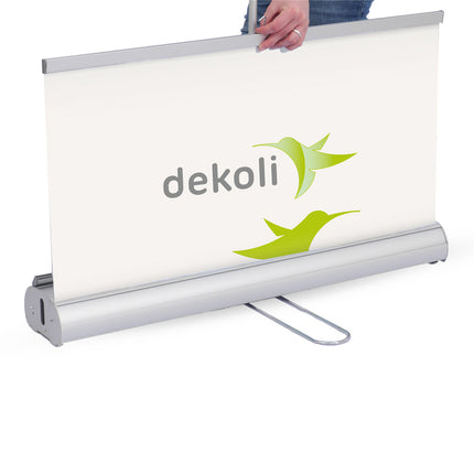 Person setzt ein Roll-Up Banner von dekolli mit grünem Logo auf einer silbernen Trägerbasis auf, ideal für sichtbare und tragbare Werbung.
